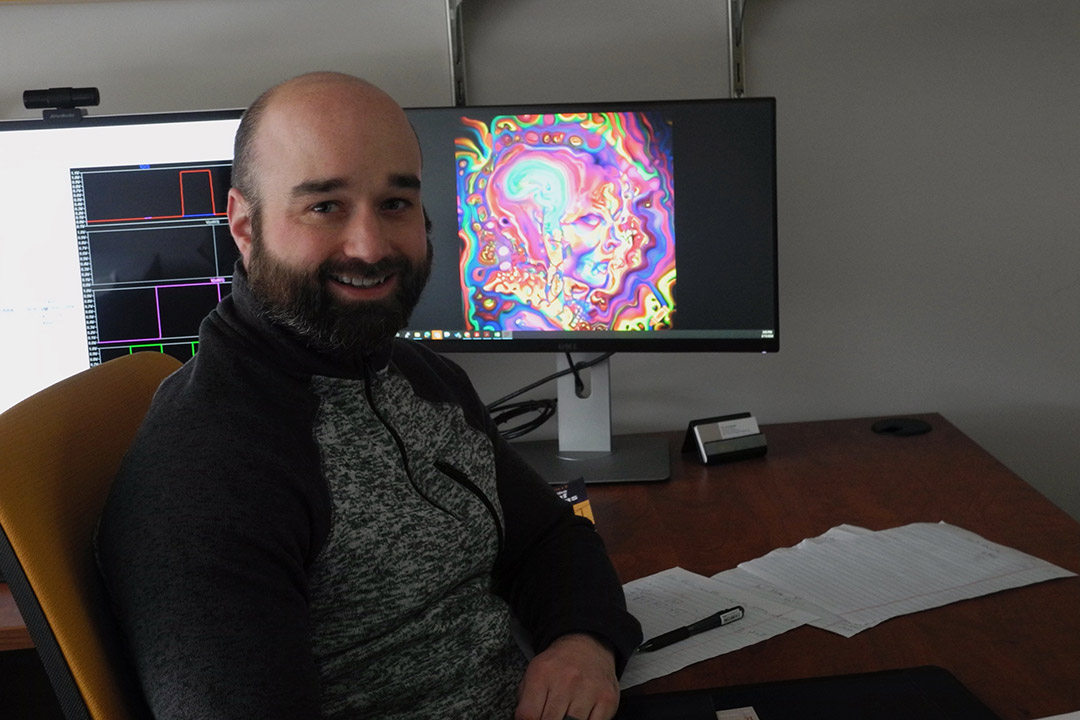Cory Merkel is shown sitting at a desk with a computer monitor showing a colorful image