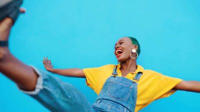 A young woman wearing overalls and a yellow t-shirt kicks one leg in the air showing excitement over the latest ASX 200 shares to hit 52-week highs