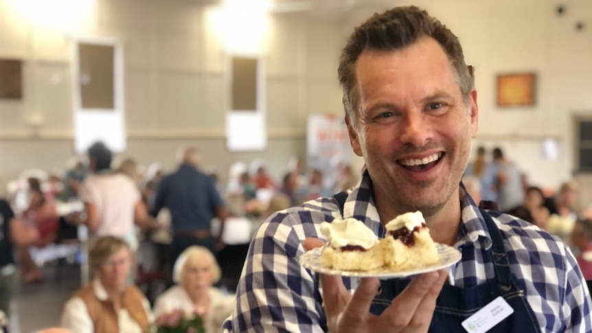 A man holds up a plate of scones with jam and cream on them and a hall full of seniors behind him.